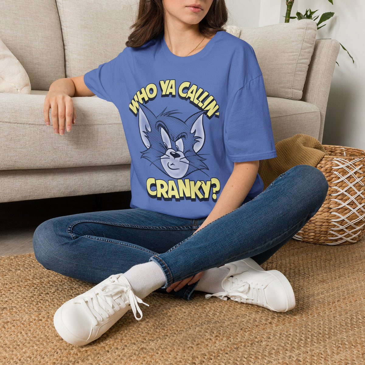 A woman sitting cross-legged on a woven rug in front of a beige sofa, wearing a blue graphic T-shirt with a cartoon cat face and the text 'WHO YA CALLIN' CRANKY?'.