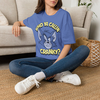 A woman sitting cross-legged on a woven rug in front of a beige sofa, wearing a blue graphic T-shirt with a cartoon cat face and the text 'WHO YA CALLIN' CRANKY?'.