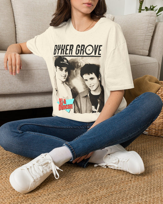 A woman sitting cross-legged on a woven rug in front of a light gray sofa, wearing an oversized cream graphic T-shirt with 'BYKER GROVE' and a black-and-white photo print, blue jeans, and white sneakers.