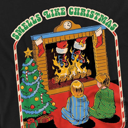 A graphic print on a black t-shirt featuring a retro Christmas scene with two children sitting in front of a burning fireplace, a decorated Christmas tree, and stockings. The text 'SMELLS LIKE CHRISTMAS' is written in green arched letters at the top.