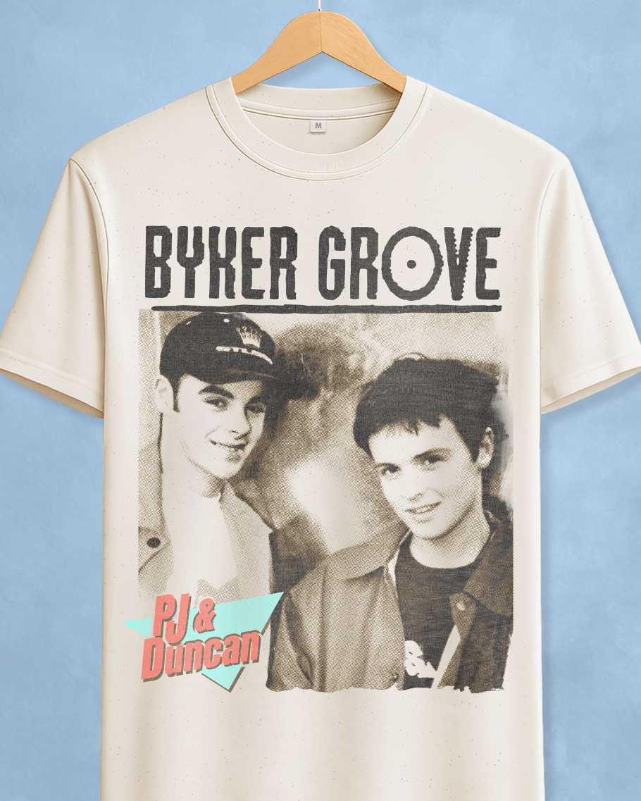 An off-white graphic t-shirt hanging on a wooden hanger against a light blue background. The shirt features a black graphic print of the text 'BYKER GROVE' above a black-and-white photo of two men, with a pink and teal 'PJ & Duncan' logo in the lower-left corner of the graphic.
