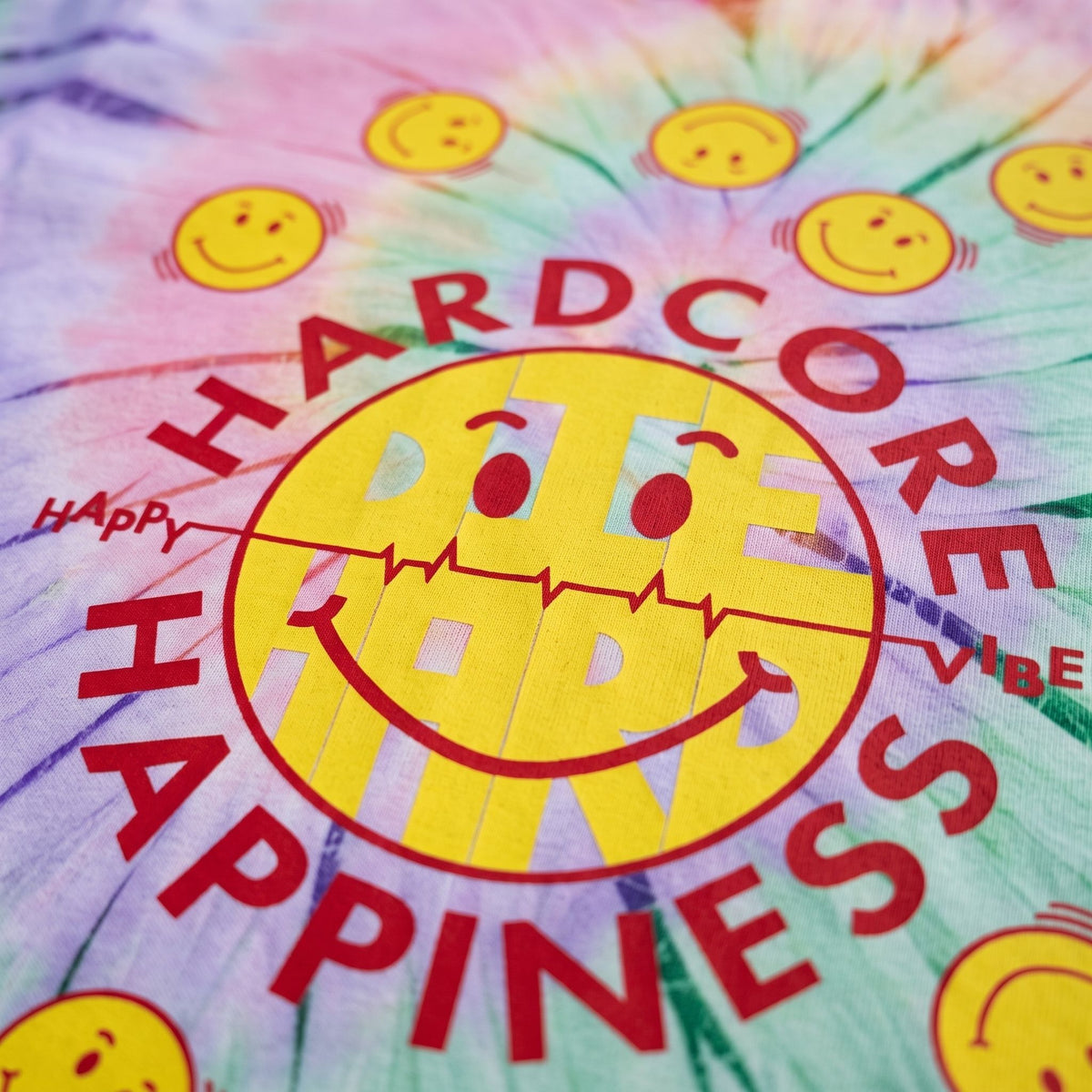 A close-up of a tie-dye fabric featuring a large yellow smiley face with a red heartbeat line across its mouth, surrounded by the words 'HARDCORE HAPPINESS' in red. Smiley faces are repeated around the circular design on a pastel tie-dye background.