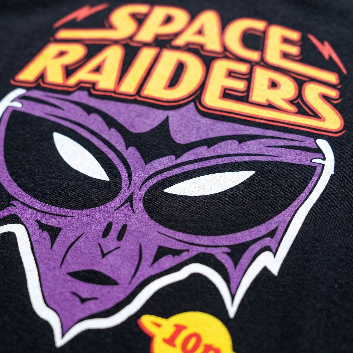 Close-up of a black graphic t-shirt featuring a stylized purple alien head with white eyes and the words 'SPACE RAIDERS' in orange and yellow text.