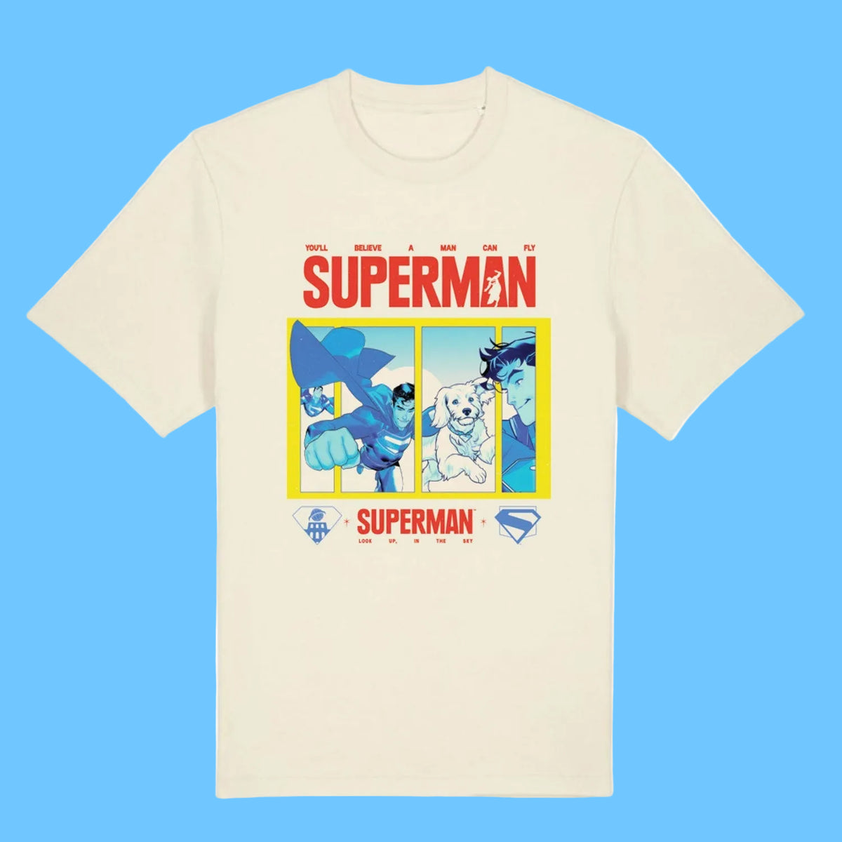 An off-white short-sleeve crewneck t-shirt featuring a colorful graphic print of Superman and other DC characters under the word 'SUPERMAN' in red, with a yellow border and small DC logos at the bottom.