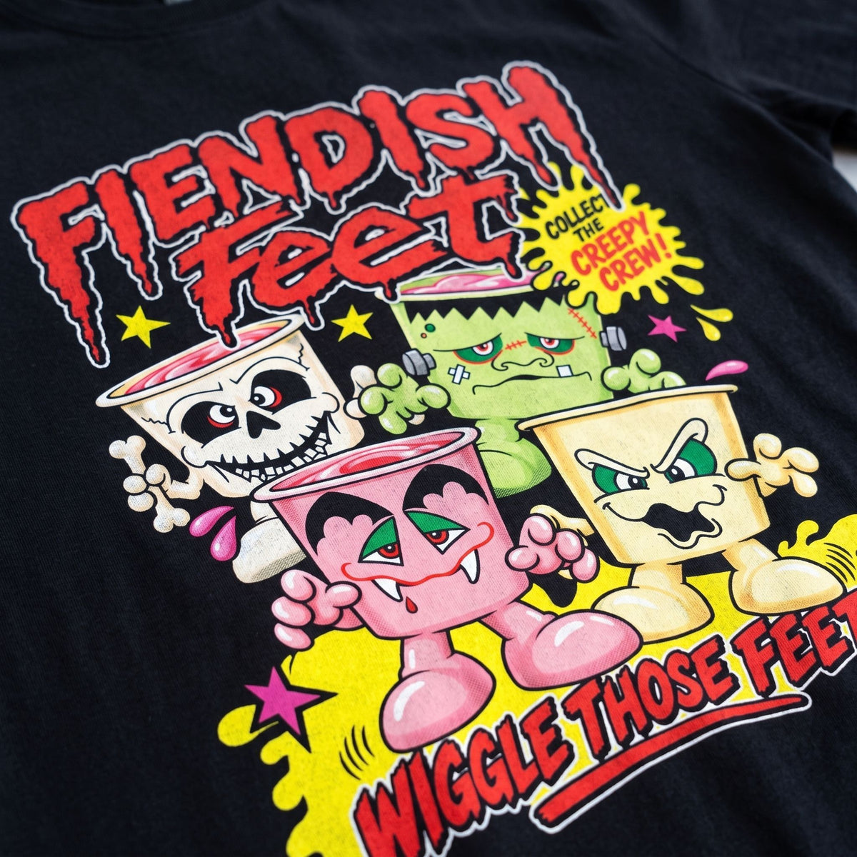 Close-up of a black graphic T-shirt featuring colorful cartoon cup characters and bold text reading 'Fiendish Feet' and 'Wiggle Those Feet.'.