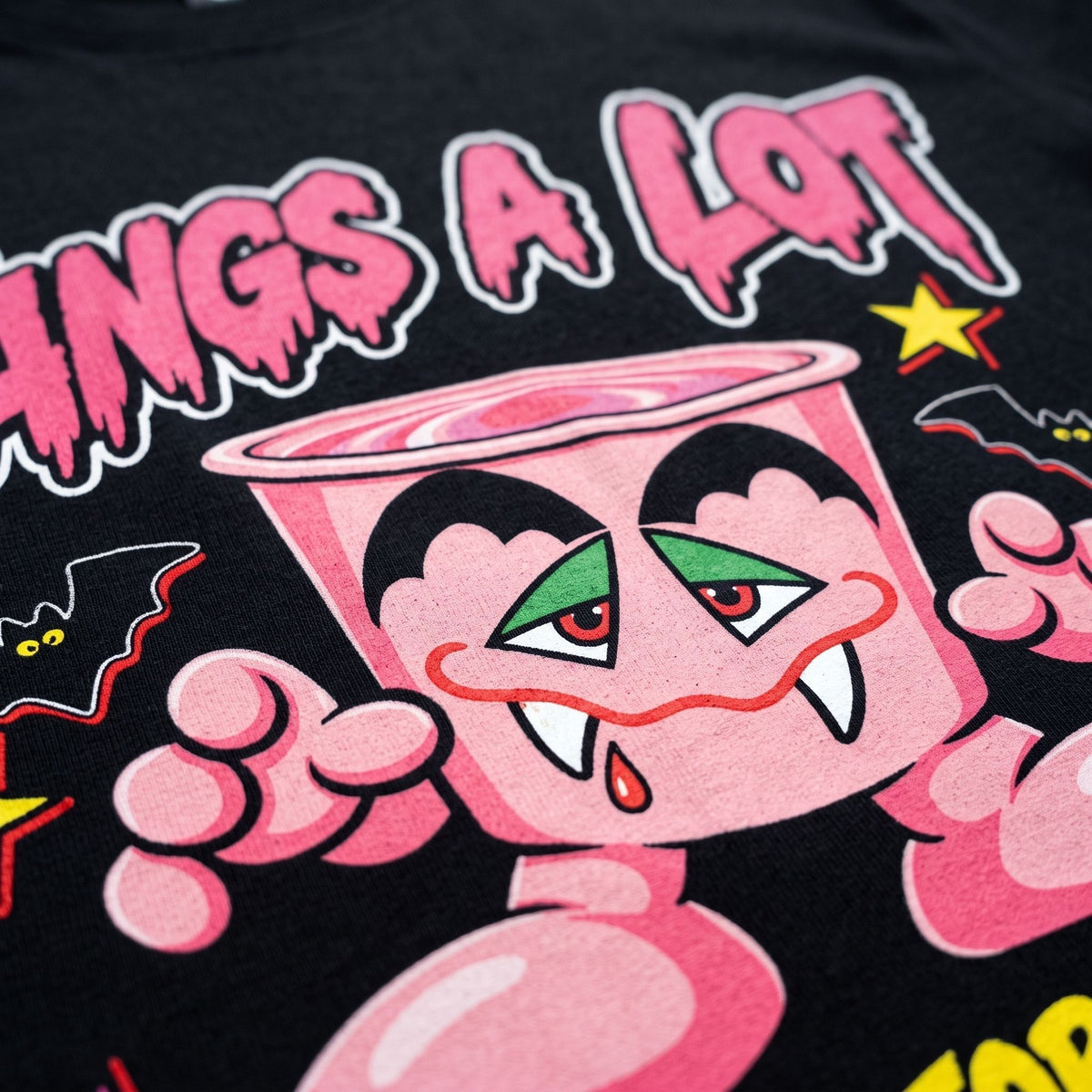 Close-up of a black graphic t-shirt featuring a pink cartoon character with green eyes, fangs, and the text 'FANGS A LOT' in dripping pink letters.