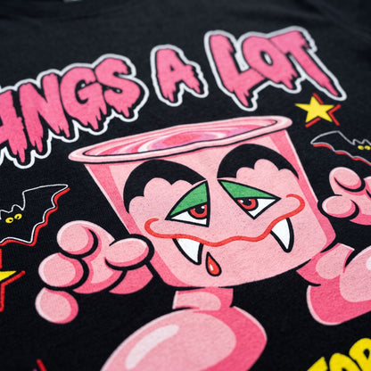 Close-up of a black graphic t-shirt featuring a pink cartoon character with green eyes, fangs, and the text 'FANGS A LOT' in dripping pink letters.