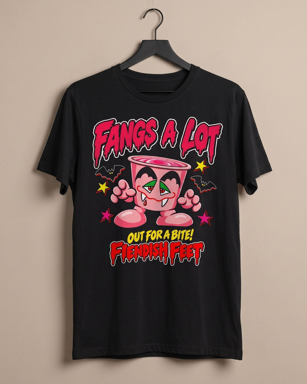 A black short-sleeve graphic t-shirt hanging on a black hanger against a solid beige wall. The shirt features a colorful cartoon graphic of a pink yoghurt pot character with green eyes and the text 'Fangs A LOT' and 'OUT FOR A BITE! Fiendish feet' in stylized red and yellow lettering.