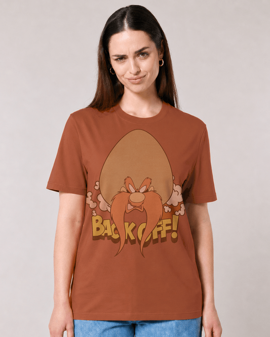 A woman modelling a brown graphic t-shirt featuring a cartoon illustration of Yosemite Sam  with the text 'BACK OFF!' in yellow. She is wearing blue jeans against a plain light gray studio background.