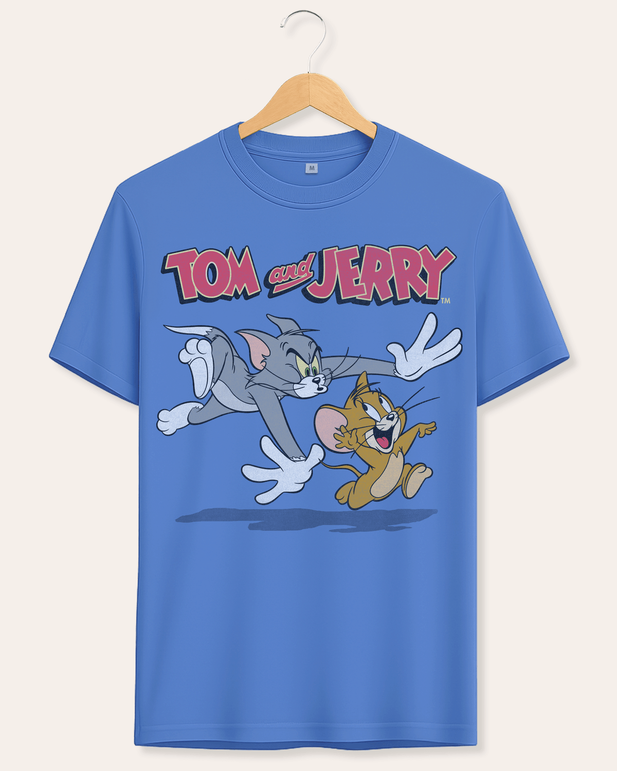 A blue short-sleeve crewneck t-shirt hanging on a wooden hanger against a light beige background. The shirt features a large graphic print of the cartoon characters Tom and Jerry running, with the 'Tom and Jerry' logo printed above them.