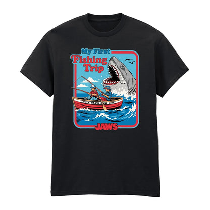 A black short-sleeve crewneck t-shirt featuring a colorful graphic print on the front. The graphic depicts a cartoon shark with an open mouth looming over a red and white fishing boat with two people on the water, with the text 'My First Fishing Trip' and 'JAWS'.