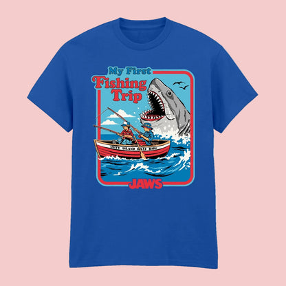 A royal blue short-sleeve t-shirt with a colorful graphic print on the front. The graphic features a cartoon scene of two people in a boat facing a giant shark, with the text 'My First Fishing Trip' and 'JAWS'.