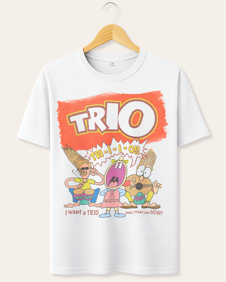A white short-sleeve crewneck t-shirt hanging on a wooden hanger against a light beige background. The shirt features a colorful cartoon graphic of three characters with long hair under a large orange 'TRIO' text banner.