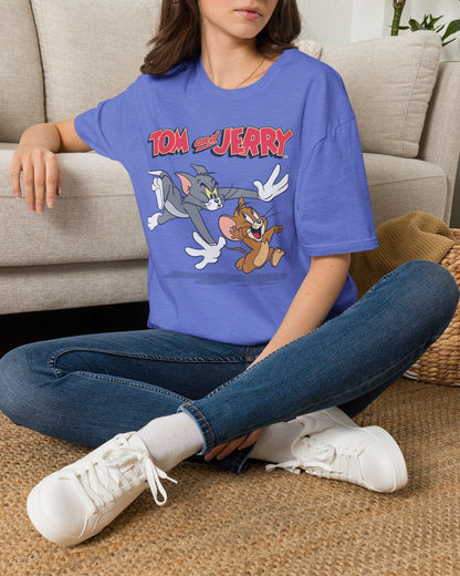 A woman sitting on a woven rug in front of a beige couch, wearing a blue graphic T-shirt with the 'Tom and Jerry' cartoon characters printed on the front, paired with blue jeans and white sneakers.