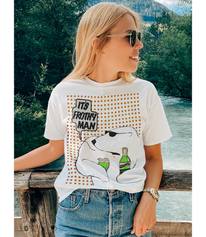 Woman wearing a t-shirt with a graphic design of a polar bear holding a fizzy pop bottle with text It's Frothy Man, standing outdoors by a river.
