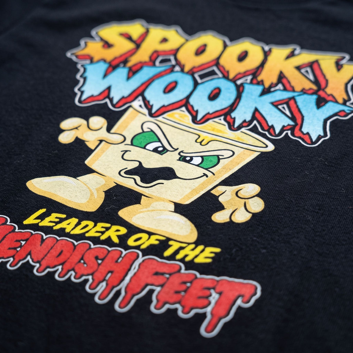Close-up of a black graphic t-shirt featuring a cartoon character and the text 'Spooky Wooky Leader of the fiendish Feet' in colorful retro lettering.