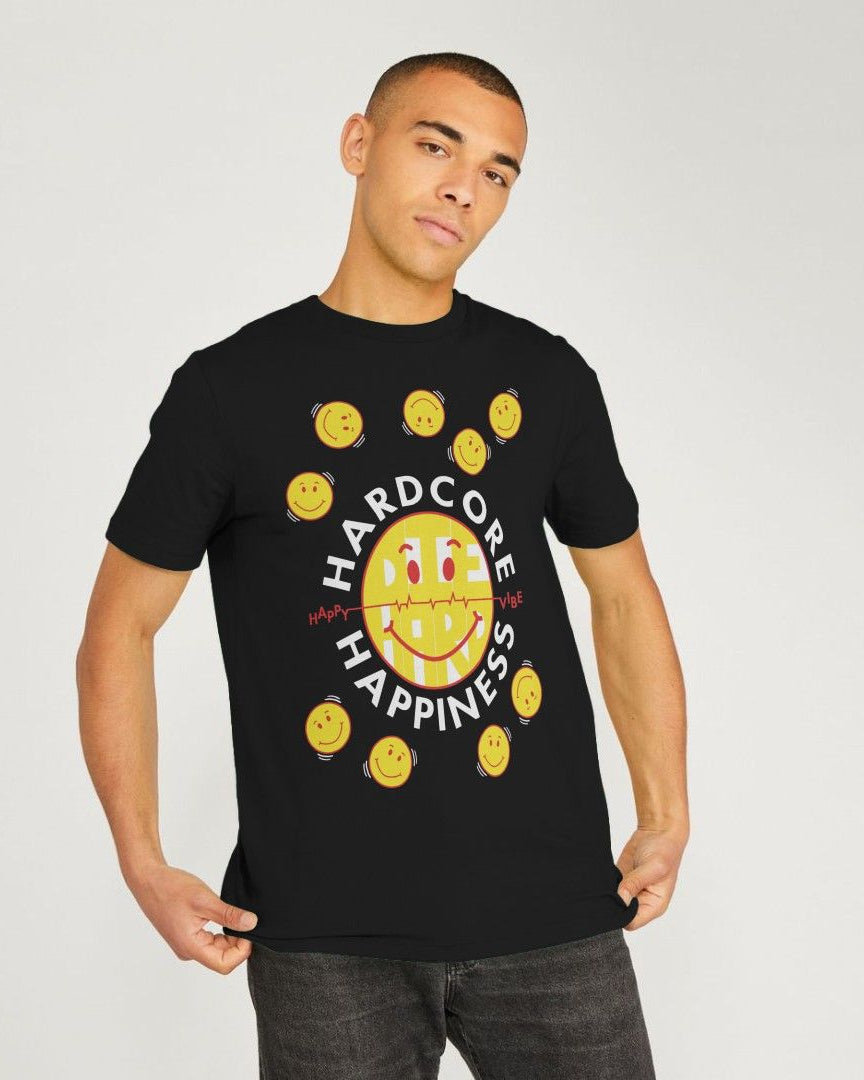 A man wearing a black short-sleeve t-shirt with a graphic print featuring a yellow smiley face, smaller yellow smiley faces around it, and the words 'HARD CORE HAPPINESS' in white. He is standing against a light gray studio background.