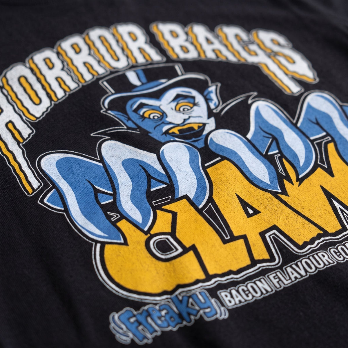 Close-up of a black graphic t-shirt featuring a blue cartoon character and bold yellow and white text that reads 'HORROR BAGS CLAWS' and '(Freaky) BACON FLAVOUR 