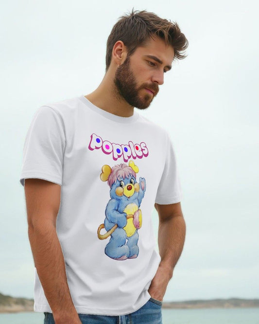 A man wearing a white graphic t-shirt with a colorful cartoon popple and the word 'poppies' printed on the front, set against a soft-focus coastal background.