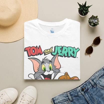 A folded white T-shirt with a colorful 'Tom and Jerry' cartoon graphic on the front, styled in a flat lay with a straw hat, white sneakers, blue jeans, sunglasses, and small potted succulents on a light beige background.