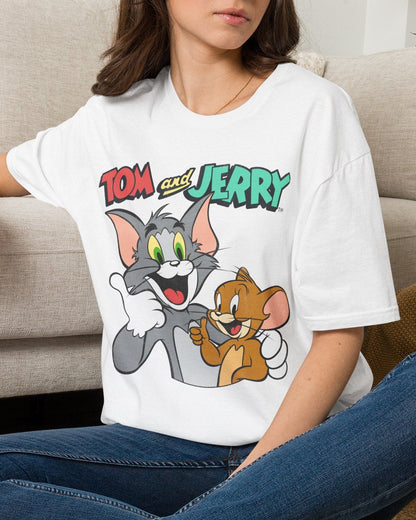 A woman sitting cross-legged on a woven rug in front of a beige sofa, wearing a white graphic T-shirt with the 'Tom and Jerry' cartoon characters printed on the front, blue jeans, and white sneakers.